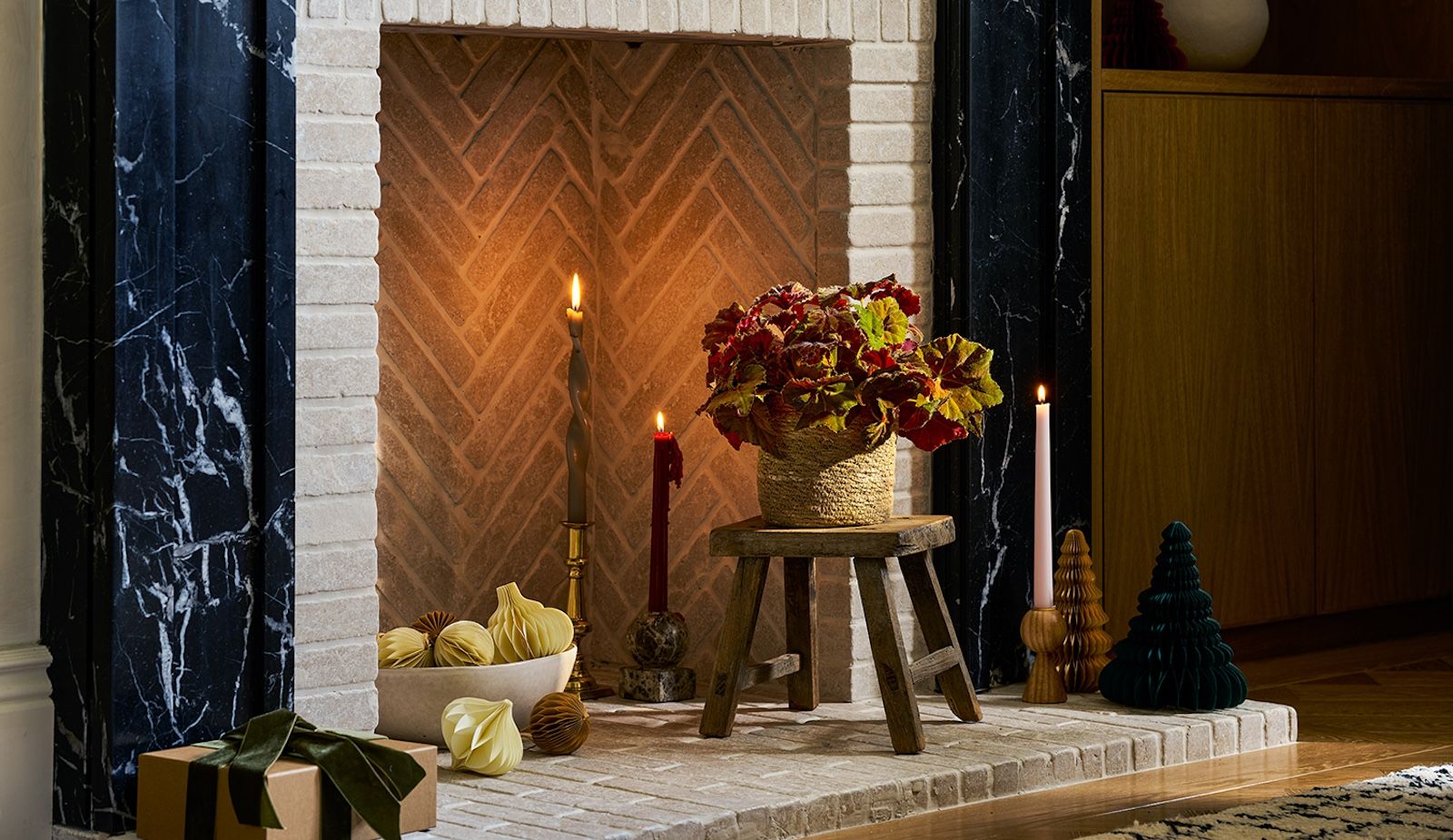 A colourful begonia in red and yellow shade sits on a wooden stool on a tiled hearth, in front of candles and Christmas decorations