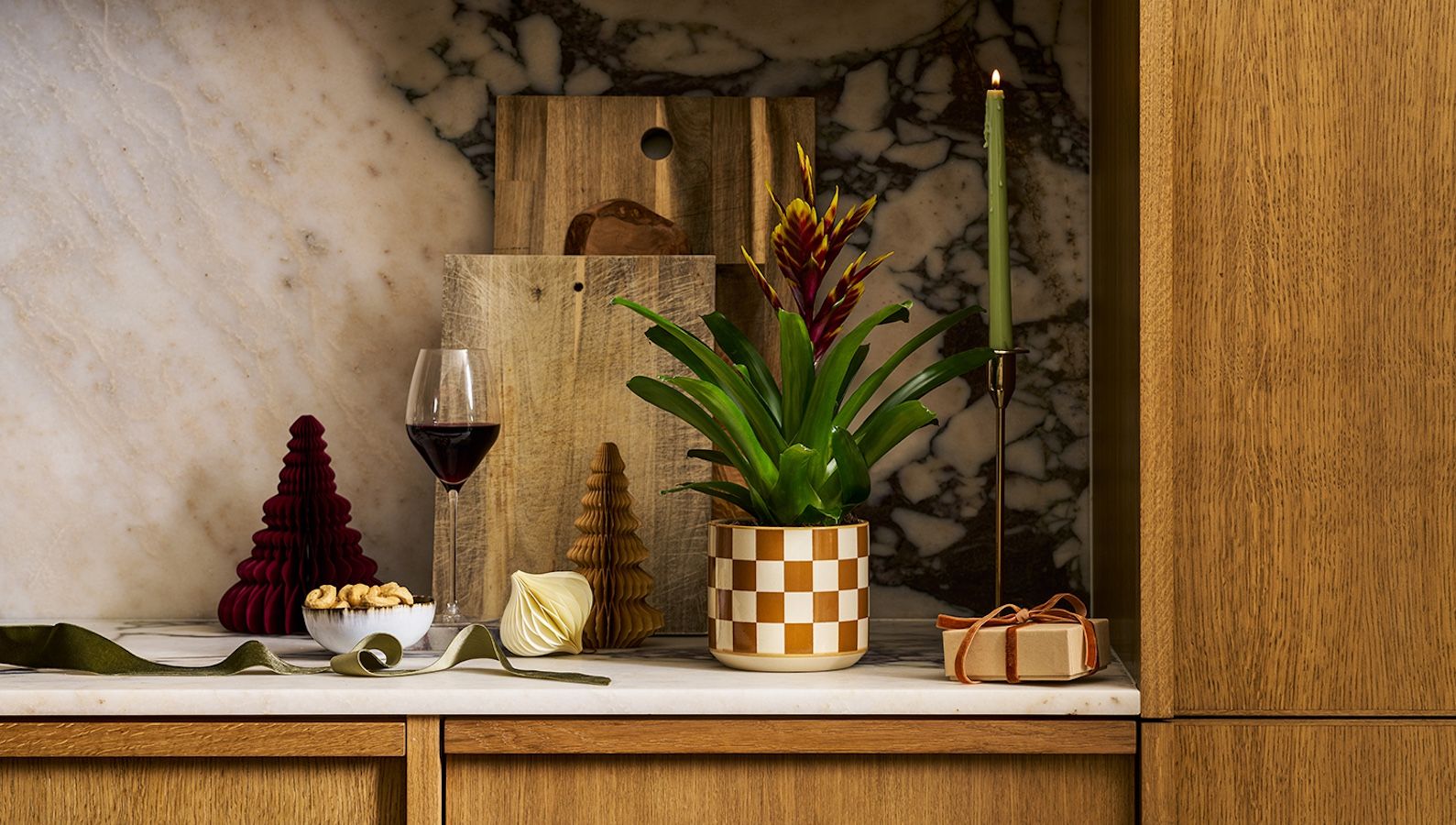 A bromeliad with a purple and yellow flower sits in a brown checked pot on a marble kitchen counter, surrounded by paper Christmas decorations, candles and a glass of wine