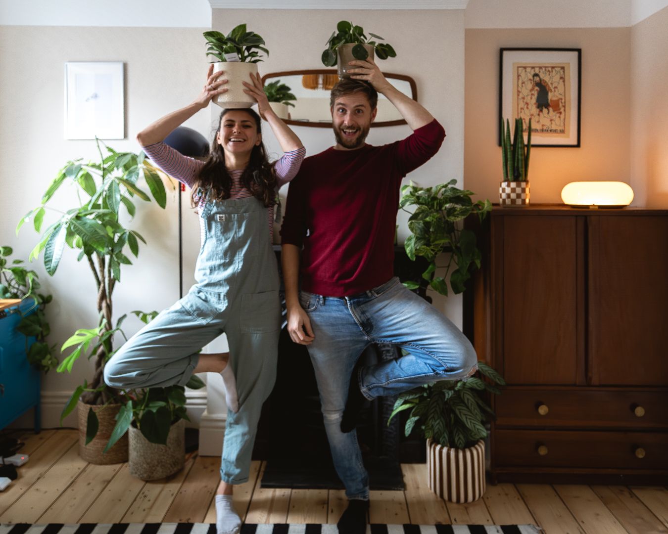 A woman wearing dungarees and a man wearing a red jumper stand in front of a fireplace with one leg resting on a knee, and their hands above their heads holding plant pots.