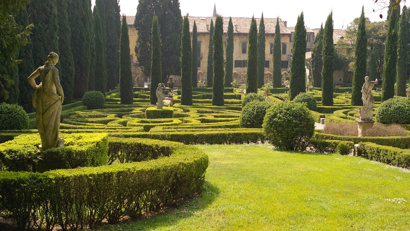 Two stone statues tower above some neat maze-like hedging. Crypress trees can be seen in the background.