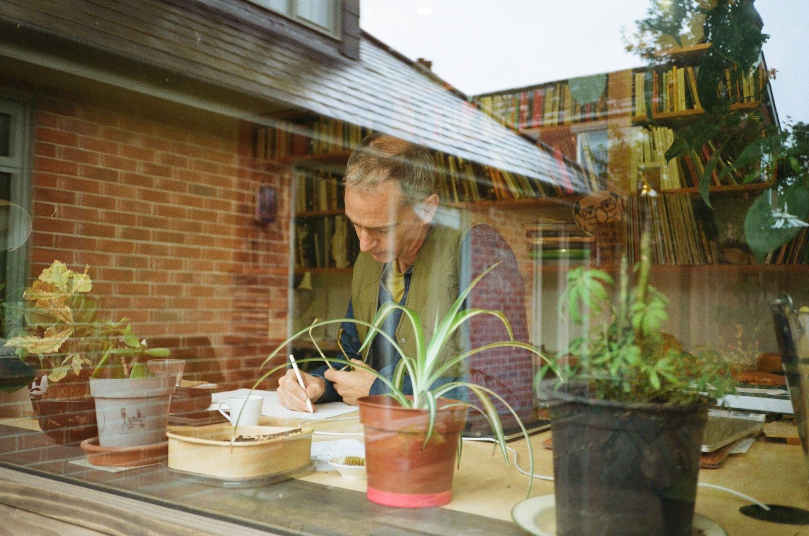 Founder of Niwaki Jake Hobson is seen writing at his desk, wearing a green quilted gilet. The photo is taken through the window, and potted plants and a bookcase can be seen in the foreground and background.