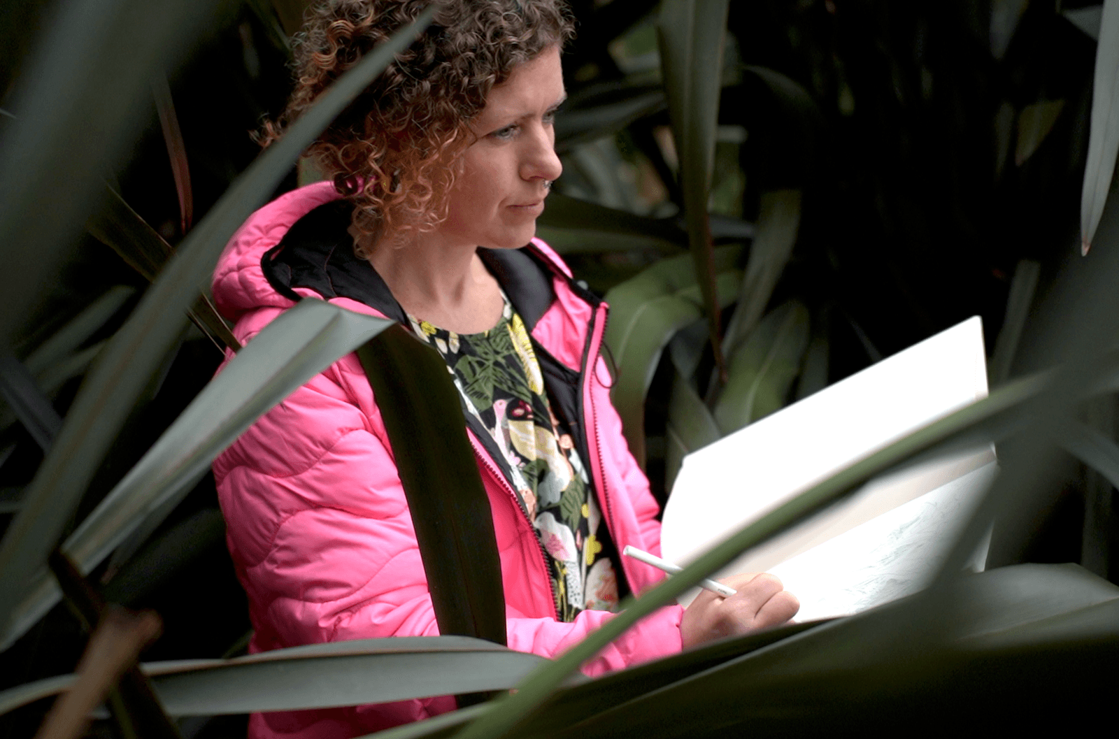 Kelly wears a neon pink jacket and sits amongst some foliage, drawing her surroundings