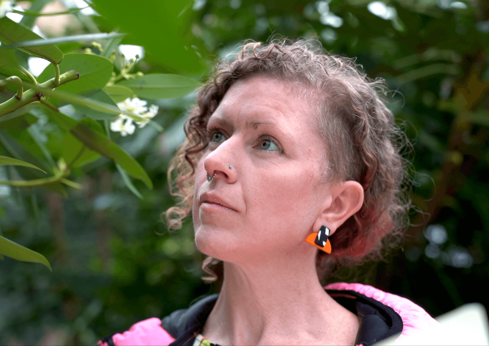 A close-up of Kelly, who is wearing orange earrings and a neon pink coat, staring up at the foliage above her