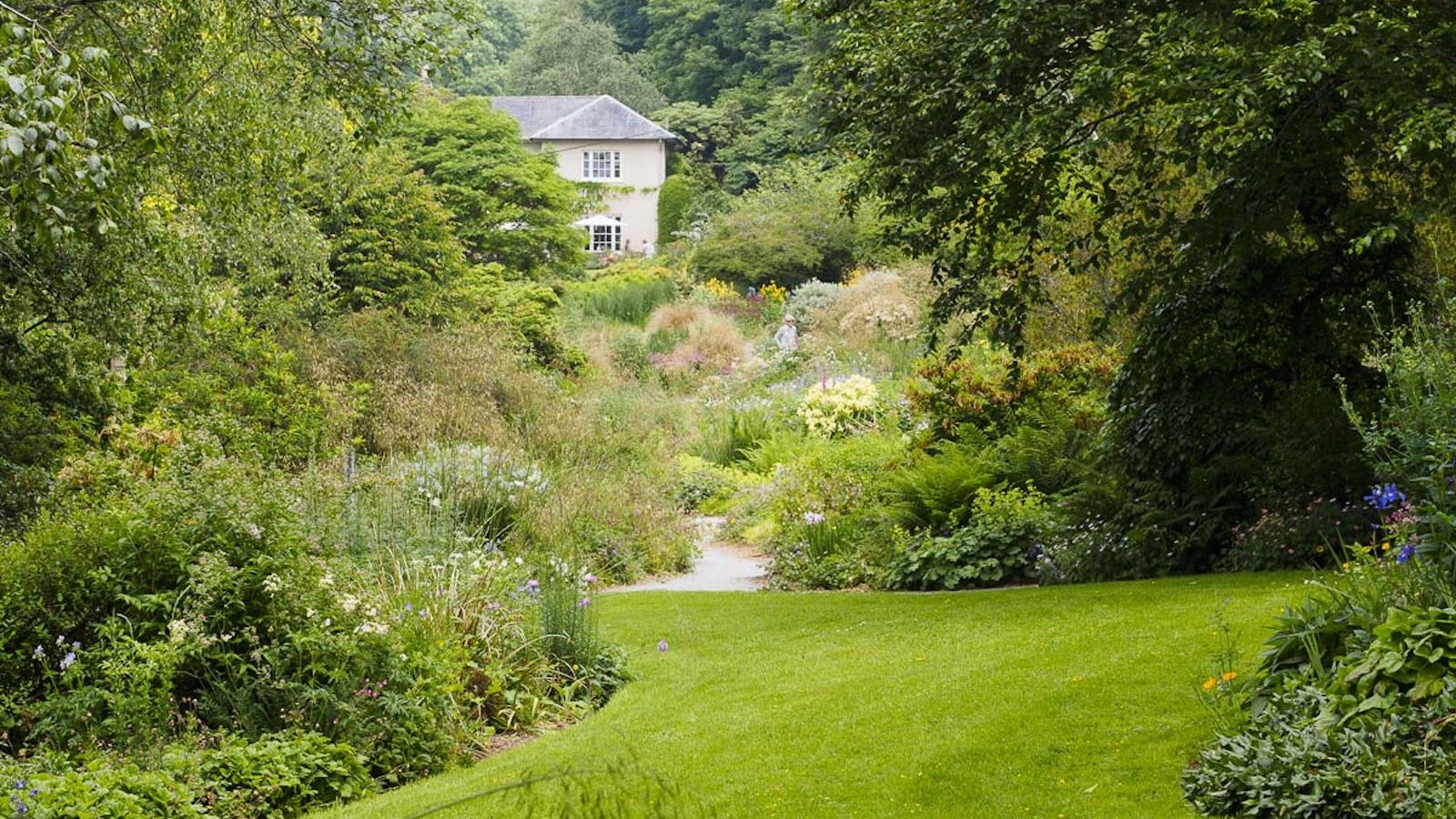 An English country garden and cottage is seen from above. Four people holding umbrellas can be seen walking down on of the garden's paths.