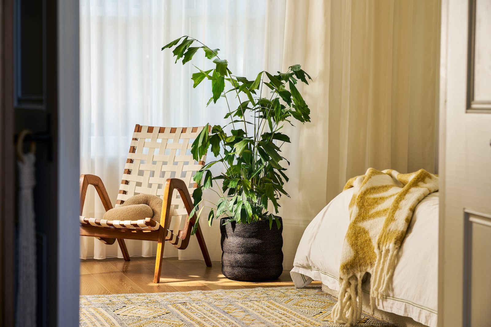 A black wave basket and fishtail palm are seen in a neutral bedroom, with a cosy yellow throw draped on the bed.